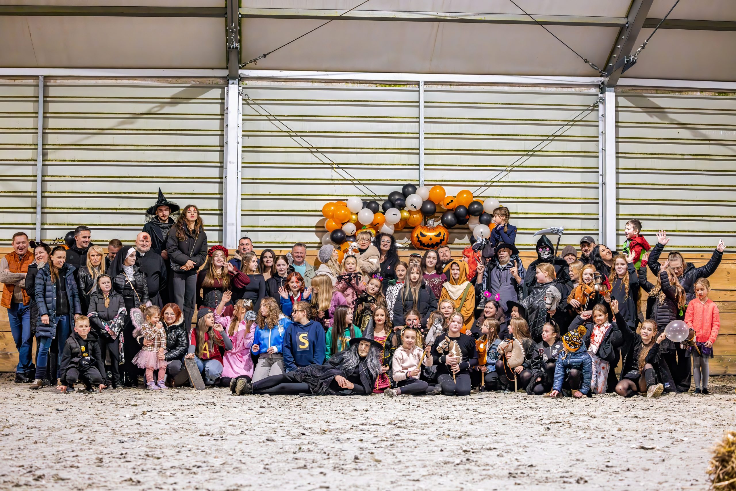 Tłum ludzi w strojach halloweenowych, głównie dzieci, przed balonami w kolorach czarnym, pomarańczowym i białym.